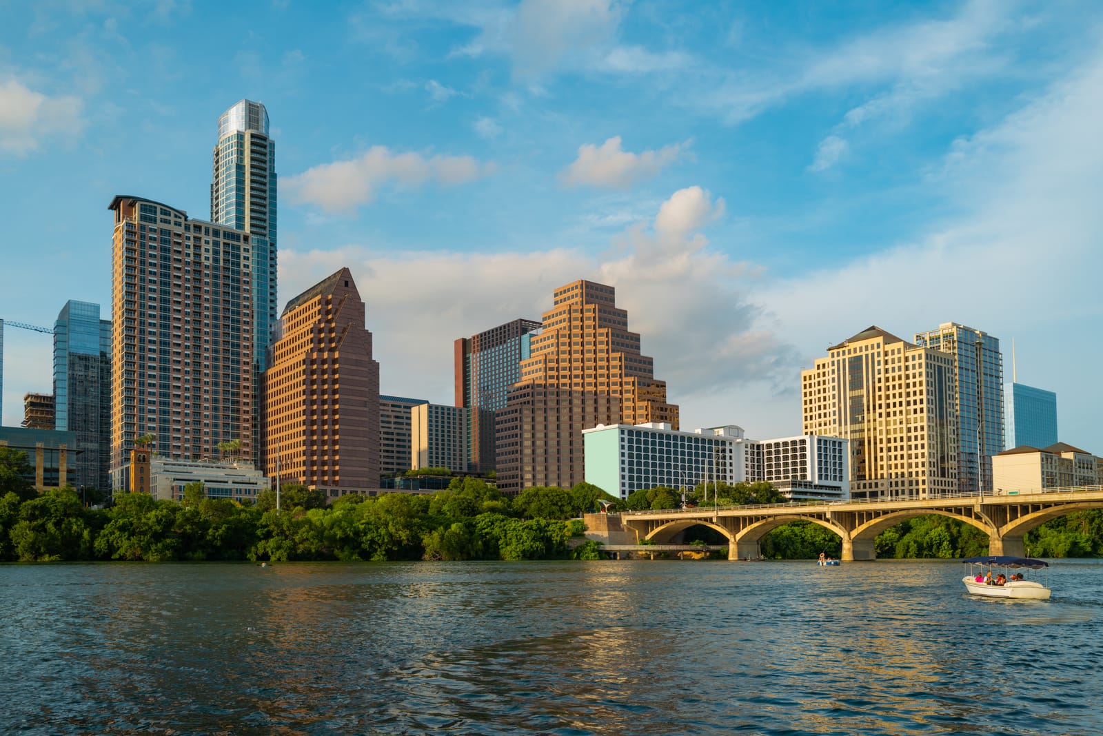 Downtown Austin towers and an arched bridge over Lady Bird Lake on a sunny day, with a boat on the water
